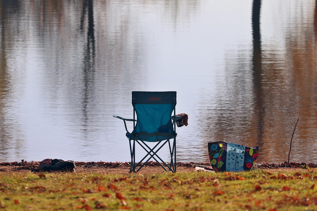 Blue camping chair by a body of water with a colorful bag nearby.